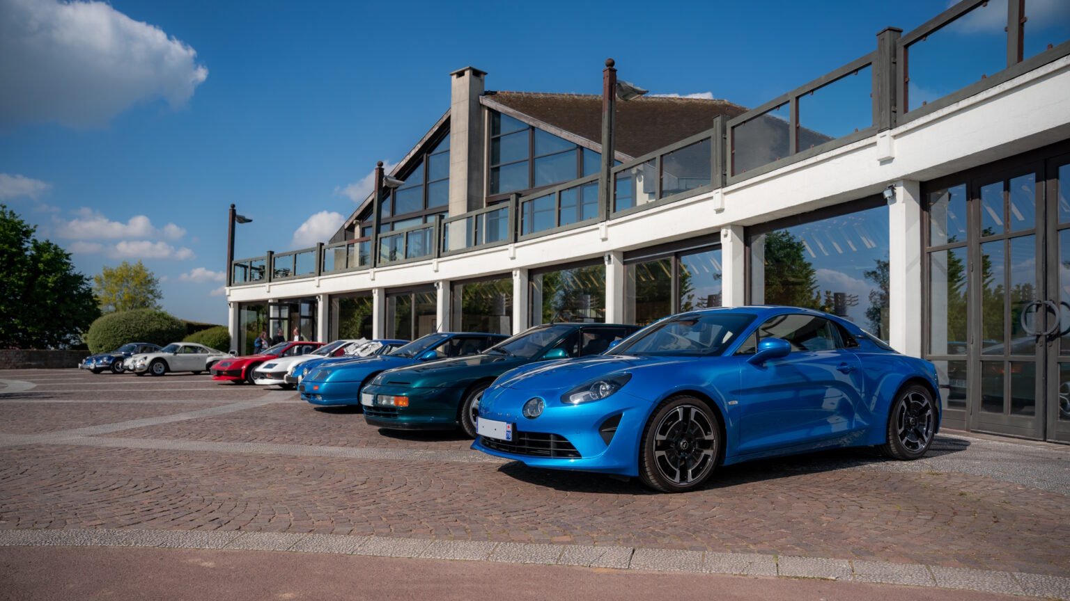 Alpine A110 et Alpine de plusieurs générations alignées devant un bâtiment vitré